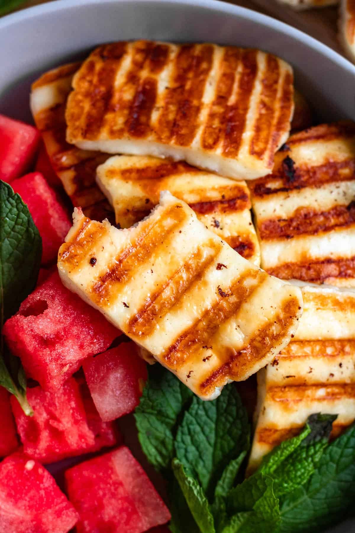A close-up image of grilled halloumi slices served with fresh watermelon cubes and mint leaves in a bowl.