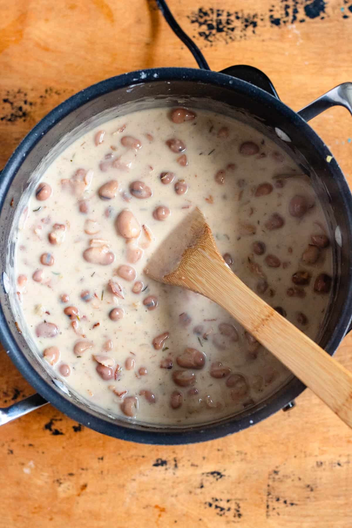 A pot of creamy beans and bacon with wooden spatula.