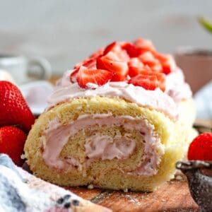 A light and fluffy strawberry Swiss roll topped with fresh strawberries sits on a wooden board.