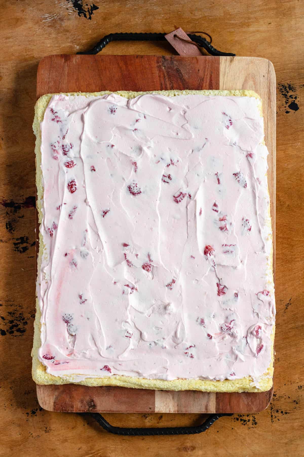 A rectangular sponge cake is evenly spread with pink strawberry cream filling, ready to be rolled into a Swiss roll.