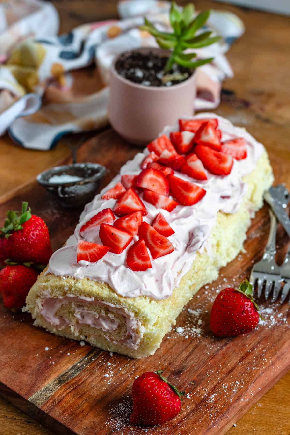 A Strawberry Swiss roll topped with fresh strawberries sits on a wooden board with fresh strawberries and forks on the side.