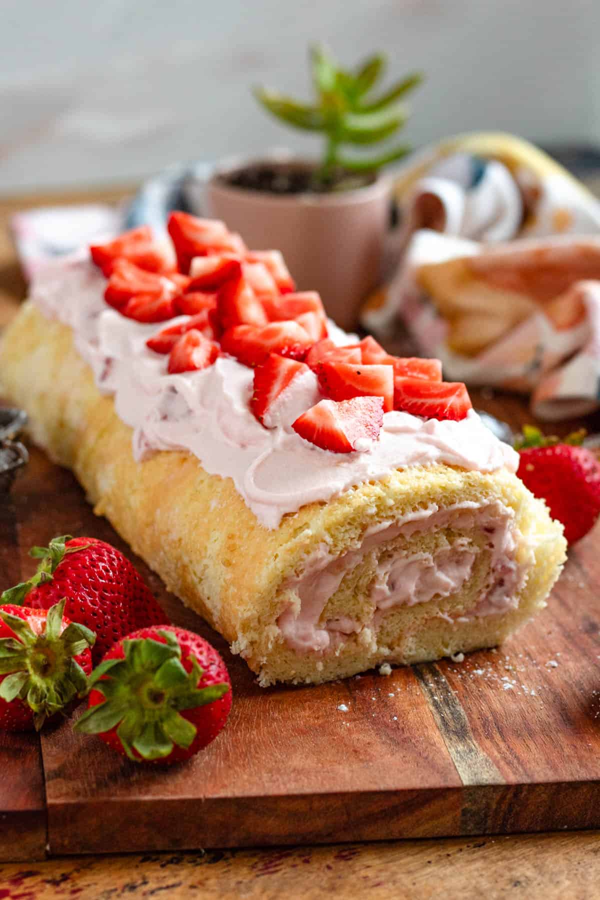 A light and fluffy strawberry Swiss roll topped with fresh strawberries sits on a wooden board with fresh strawberry pieces on the side.