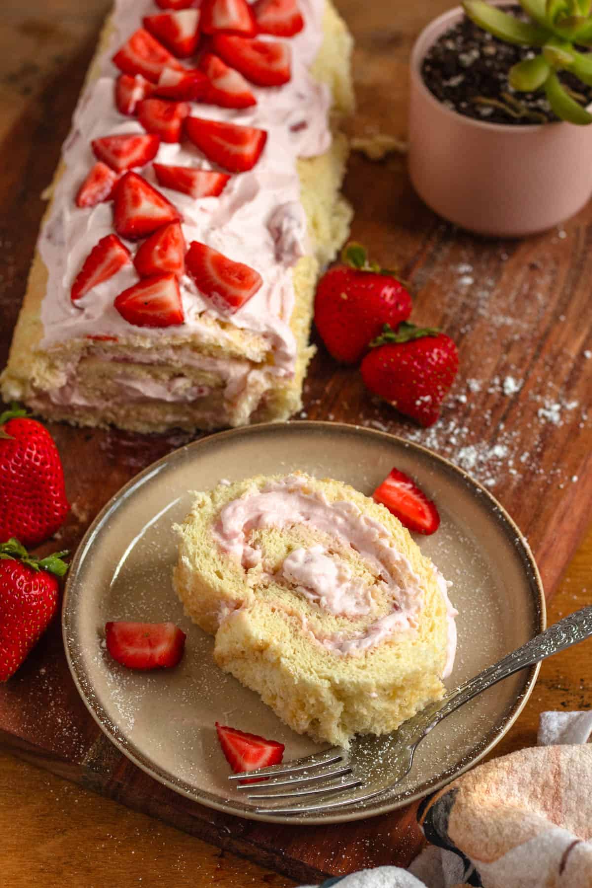 A slice of strawberry Swiss roll is served on a plate, while the full, strawberry-topped roll rests in the background on a wooden board.