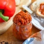 A jar of vibrant Ajvar recipe sits on a rustic table beside fresh bread and a whole red bell pepper.