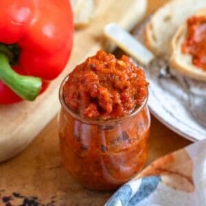 A jar of vibrant Ajvar recipe sits on a rustic table beside fresh bread and a whole red bell pepper.