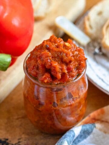 A jar of vibrant Ajvar recipe sits on a rustic table beside fresh bread and a whole red bell pepper.