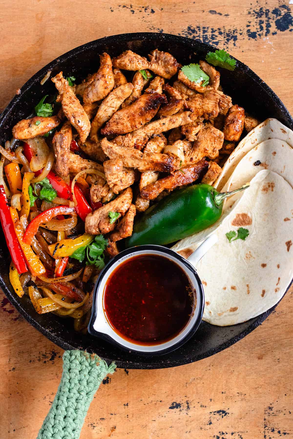 A skillet filled with seasoned chicken, sautéed peppers and onions, tortillas, a jalapeño, and a cup of rich fajita sauce.