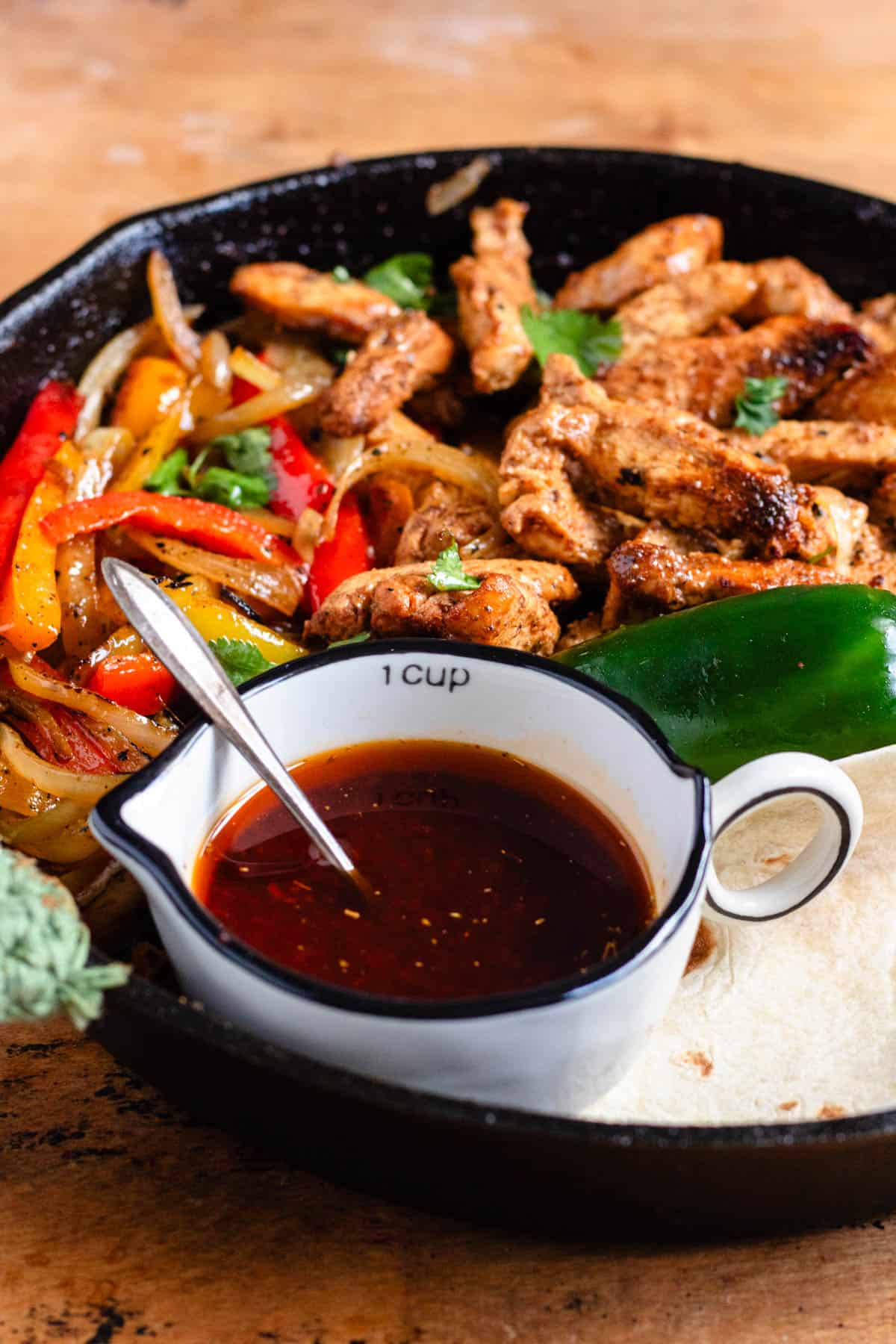 A close-up of a skillet brimming with seasoned chicken, sautéed peppers and onions, and a cup of rich fajita sauce.