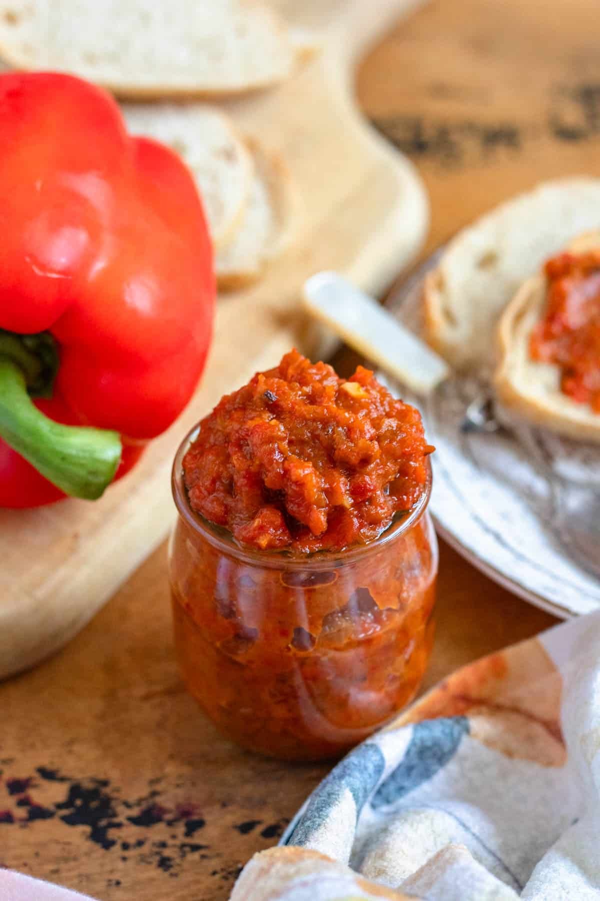 A jar of vibrant Ajvar recipe sits on a rustic table beside fresh bread and a whole red bell pepper.
