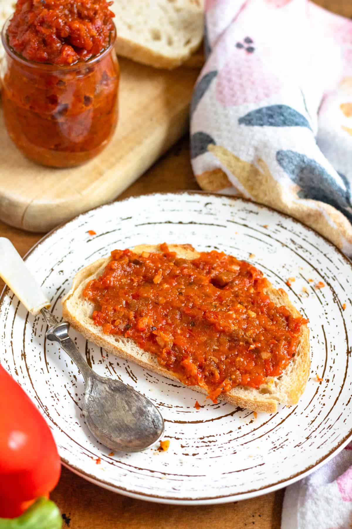 A slice of bread topped with chunky homemade ajvar recipe sits next to a jar filled with the same spread.