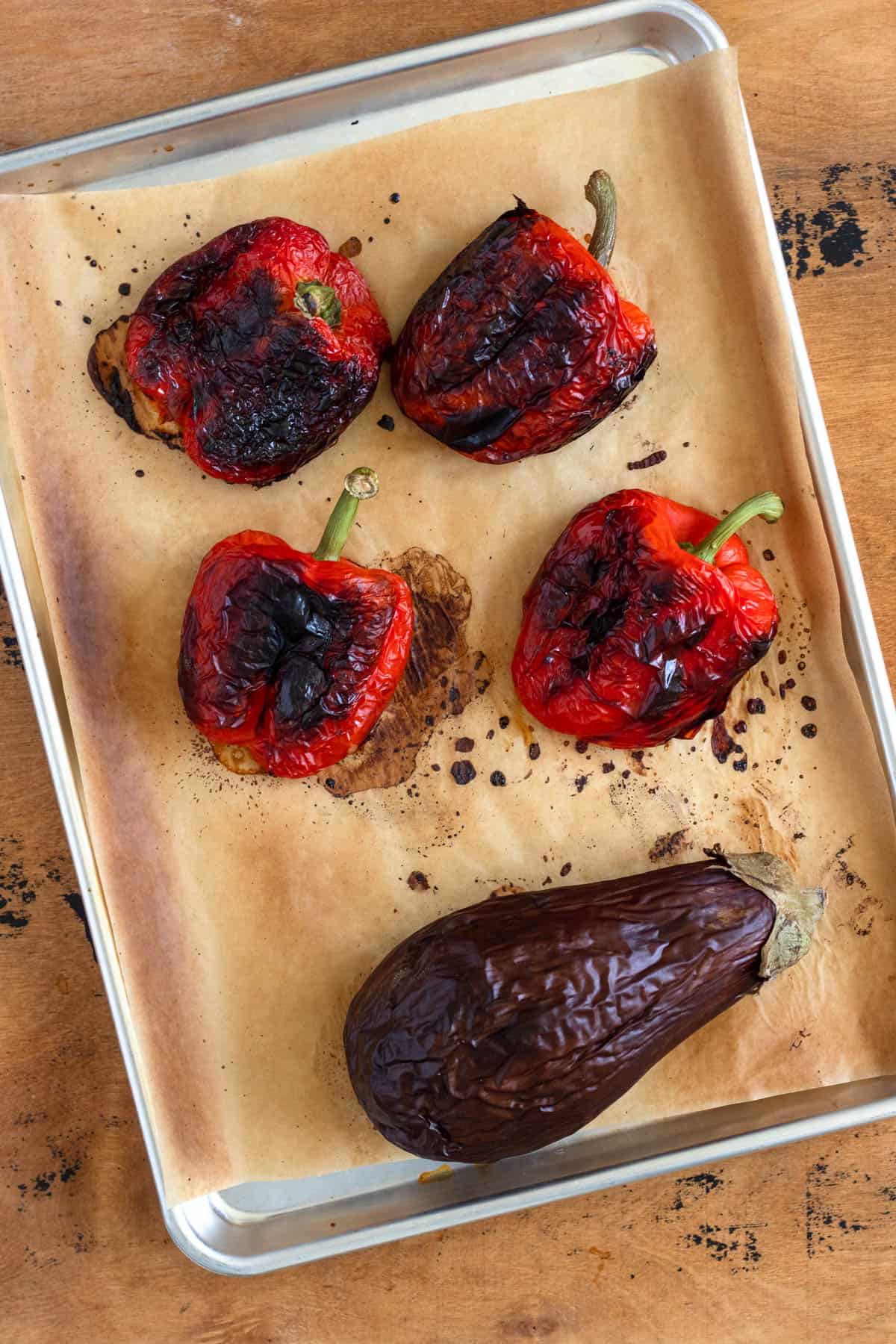 Charred red bell peppers and a roasted eggplant rest on a parchment-lined baking sheet.
