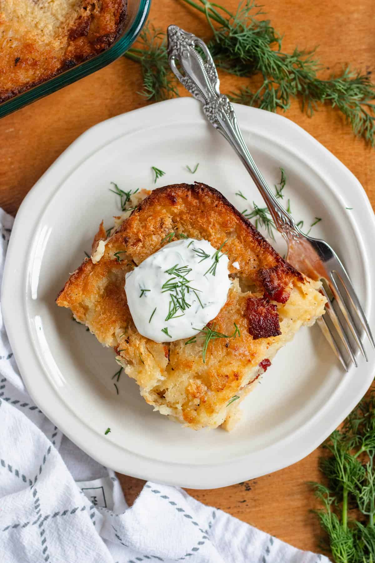 A slice of golden-brown Lithuanian potato kugelis topped with sour cream and fresh dill on a white plate with a fork.