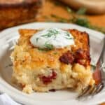 A slice of golden-brown Lithuanian potato kugelis topped with sour cream and fresh dill on a white plate with a fork.