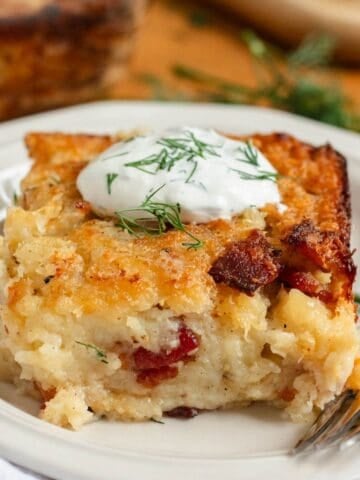 A slice of golden-brown Lithuanian potato kugelis topped with sour cream and fresh dill on a white plate with a fork.