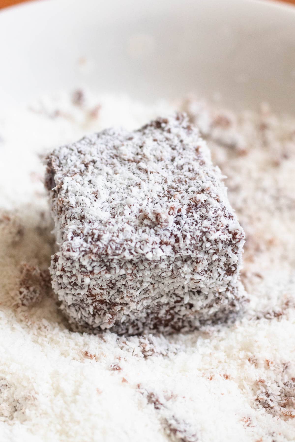 A chocolate covered piece of cake in a shallow bowl of desiccated coconut. 