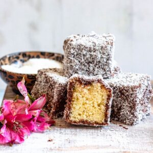 Lamingtons stacked together on a plate with bright pink flowers garnished on the side.