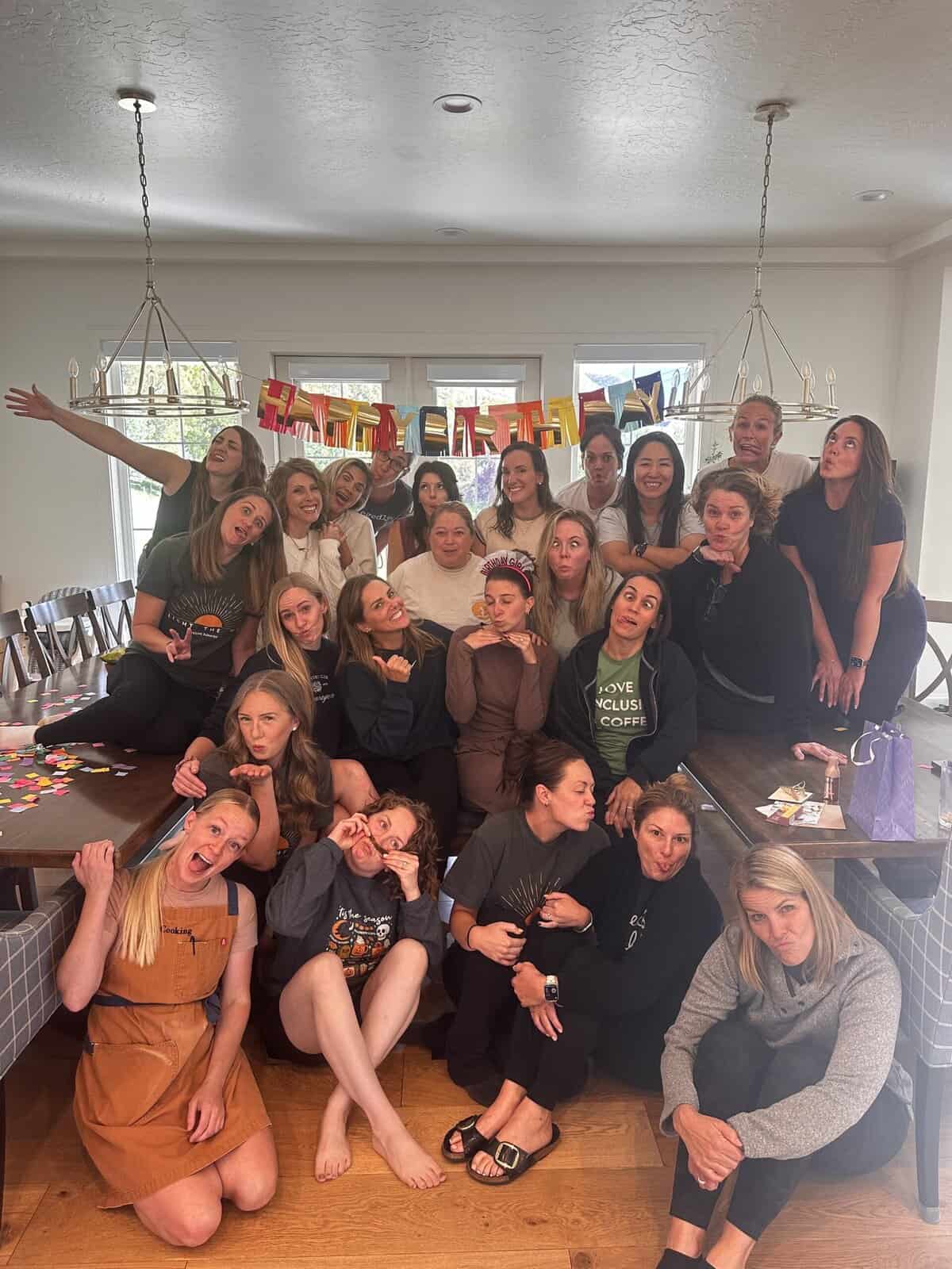 A group of women in a kitchen posing goofily with a happy birthday sign above them.