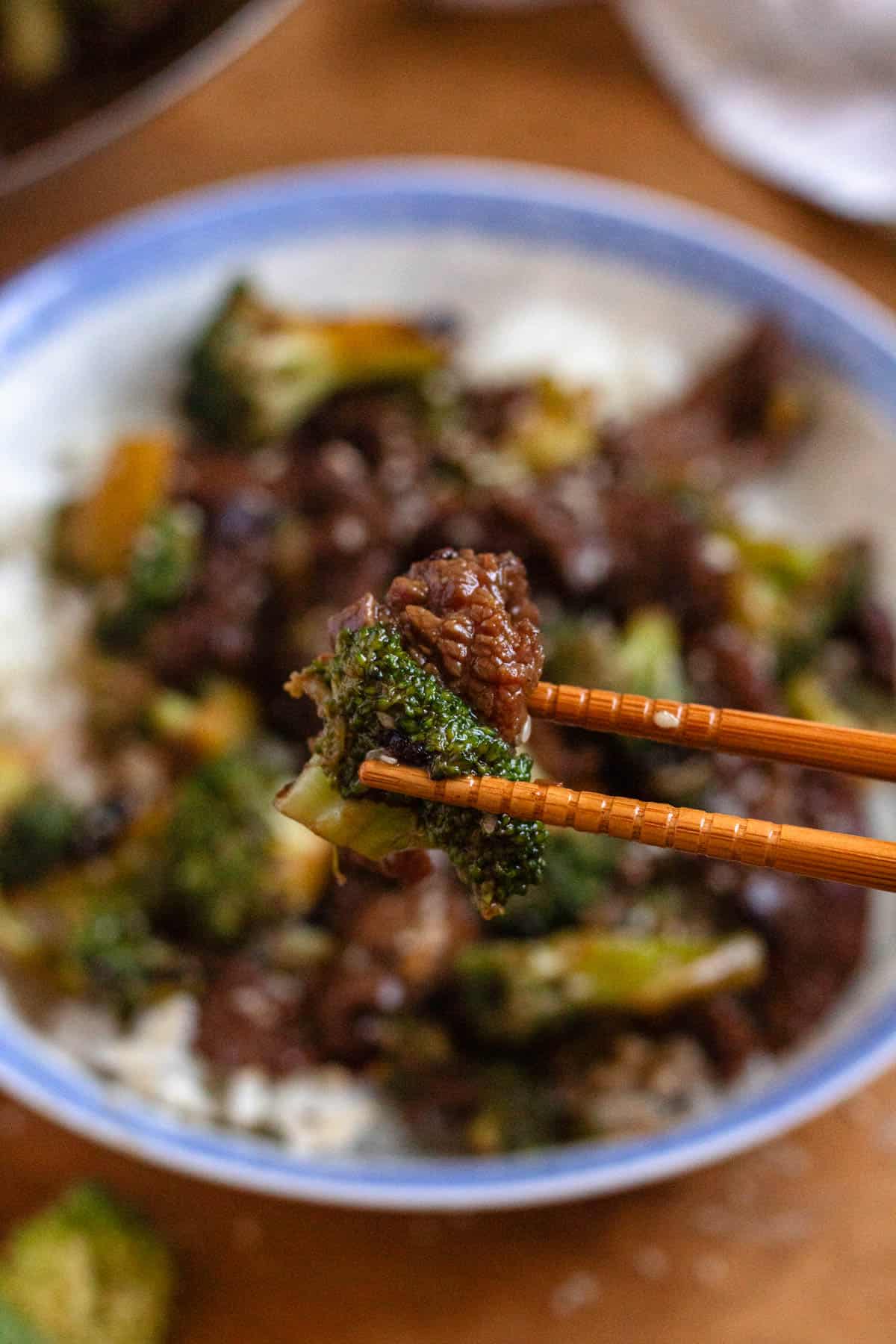 Chopsticks holding a bite of beef and broccoli over a bowl of rice.