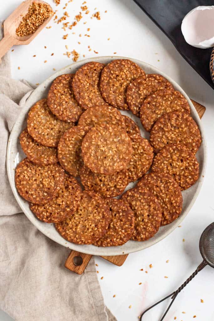 A plate filled with thin, crispy benne wafers on a light countertop with scattered sesame seeds around.
