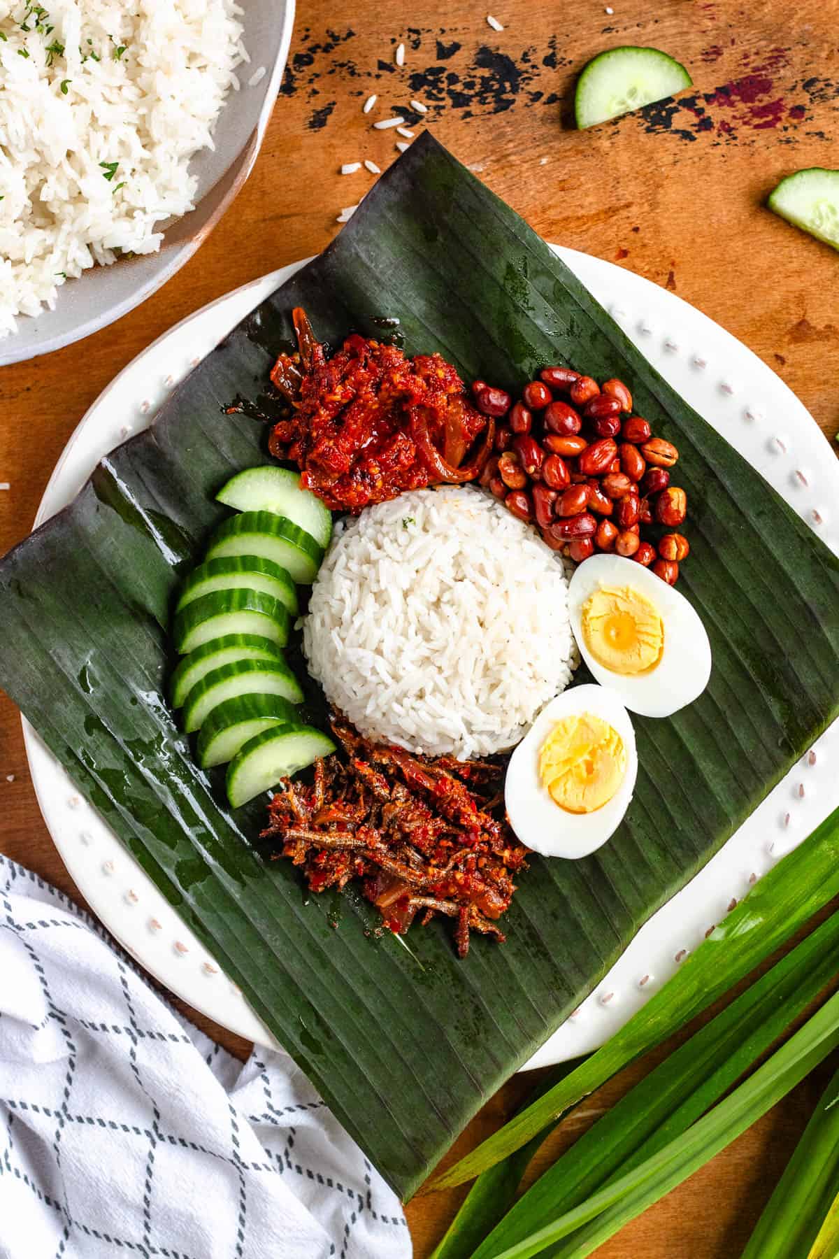A plate of Malaysian Nasi Lemak served with rice, sambal, boiled egg, peanuts, anchovies, and cucumber slices on a banana leaf.