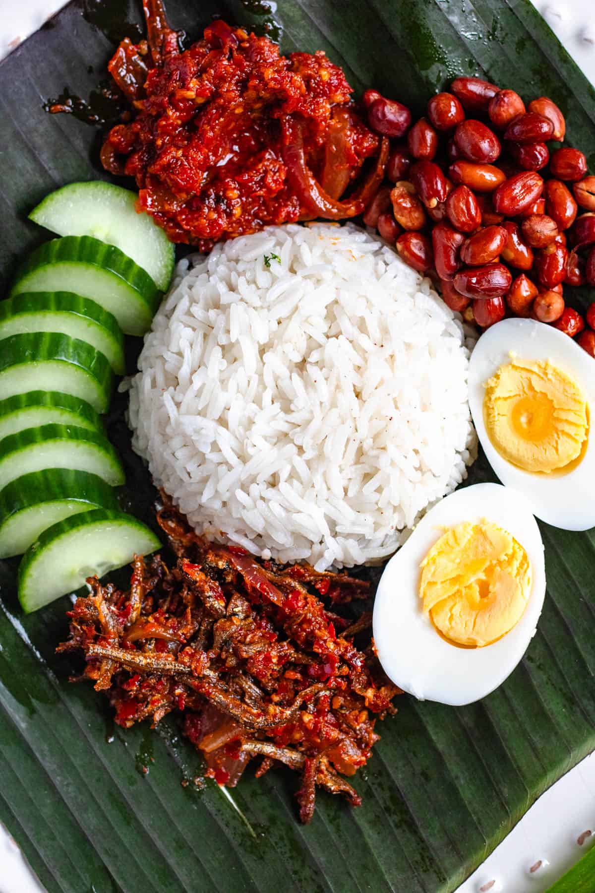 A plate of Malaysian Nasi Lemak served with rice, sambal, boiled egg, peanuts, anchovies, and cucumber slices on a banana leaf.