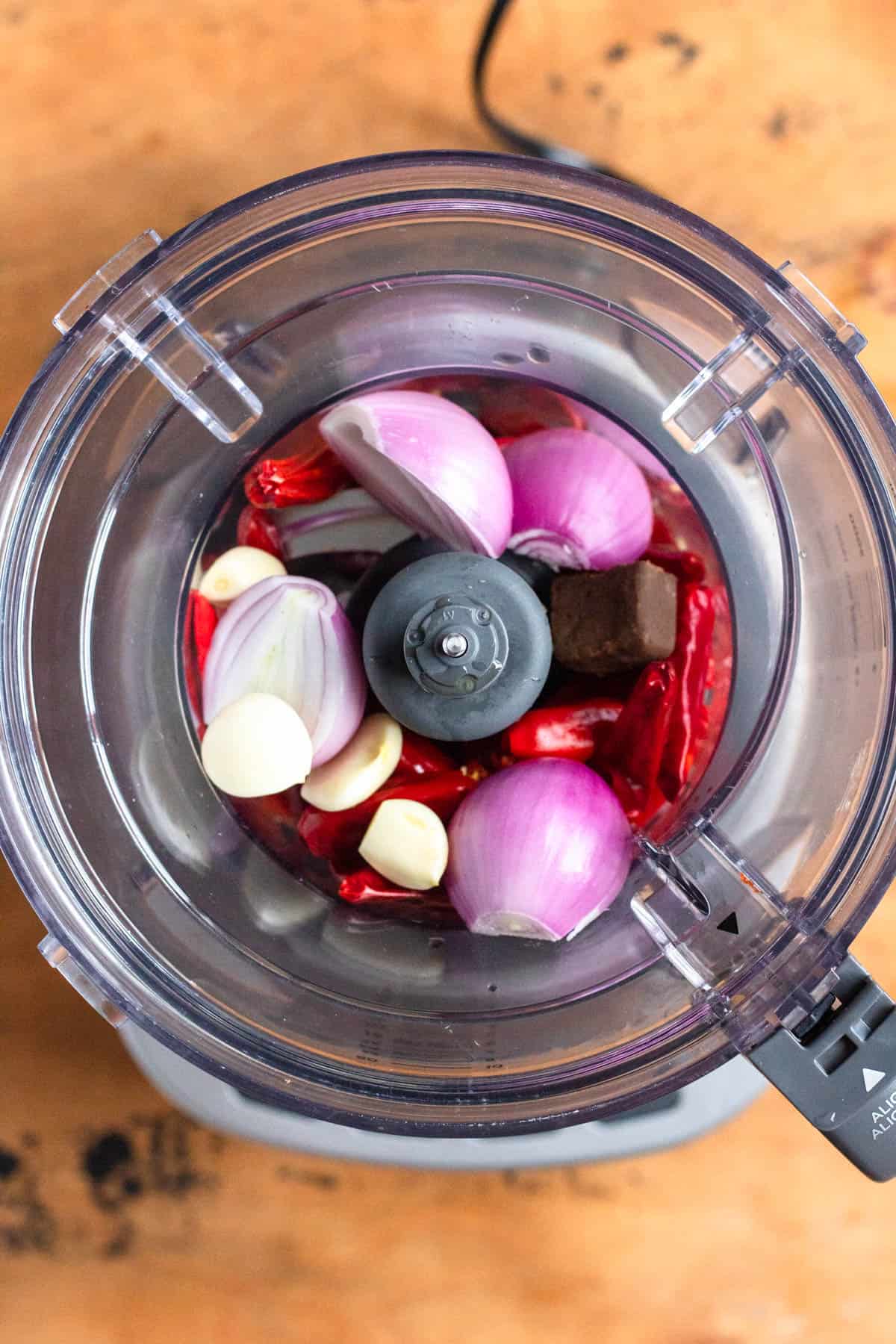 Ingredients including rehydrated chilies, shrimp paste, shallots, garlic, and water placed in a food processor.