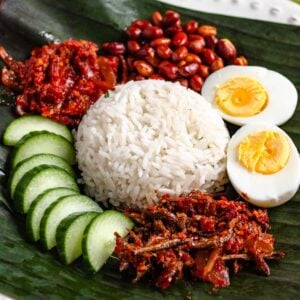 A plate of Malaysian Nasi Lemak served with rice, sambal, boiled egg, peanuts, anchovies, and cucumber slices on a banana leaf.
