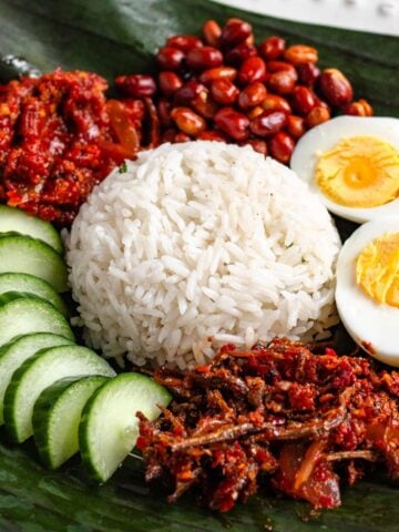 A plate of Malaysian Nasi Lemak served with rice, sambal, boiled egg, peanuts, anchovies, and cucumber slices on a banana leaf.
