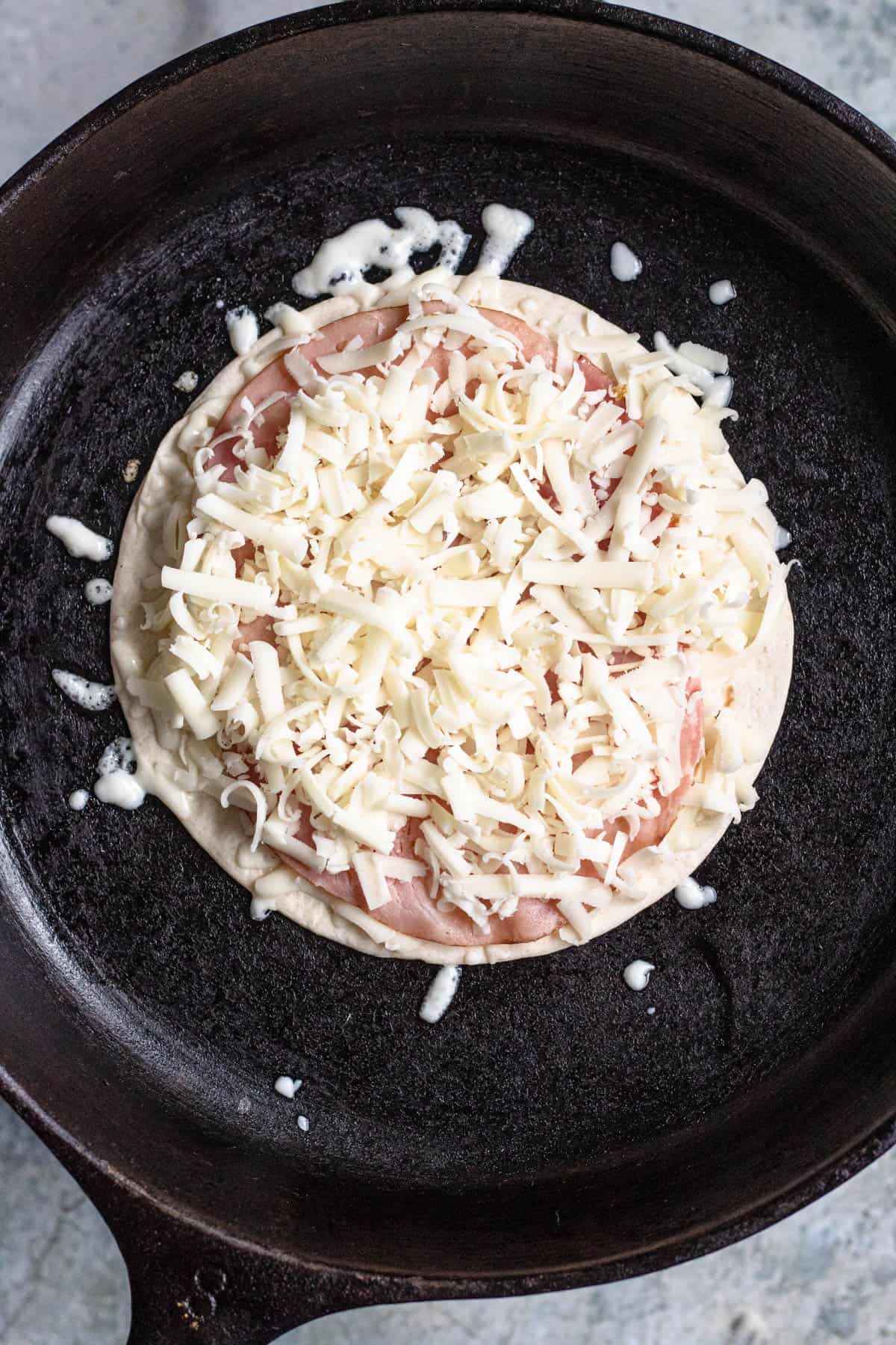 A flour tortilla topped with ham and cheese warming in a cast iron skillet to prepare sincronizadas.