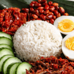 Pinterest image of a close-up view of a plate of Malaysian Nasi Lemak served with rice, sambal, boiled egg, peanuts, anchovies, and cucumber slices on a banana leaf.