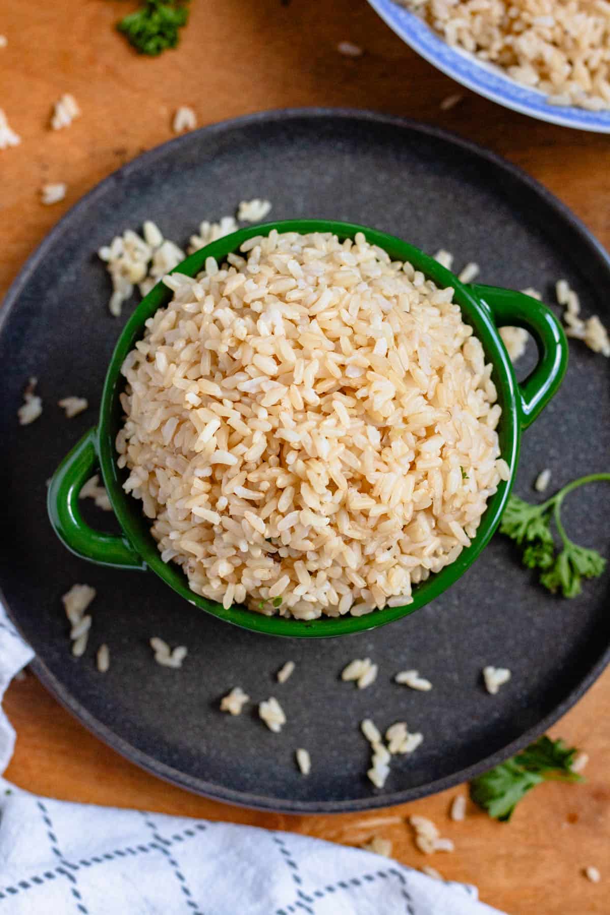 A bowl of cooked brown rice sits on a dark plate with a few scattered grains and fresh herbs around it.
