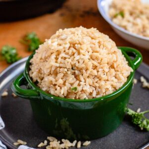 A green bowl filled with cooked brown rice sits on a green bowl with scattered grains and herbs around it.
