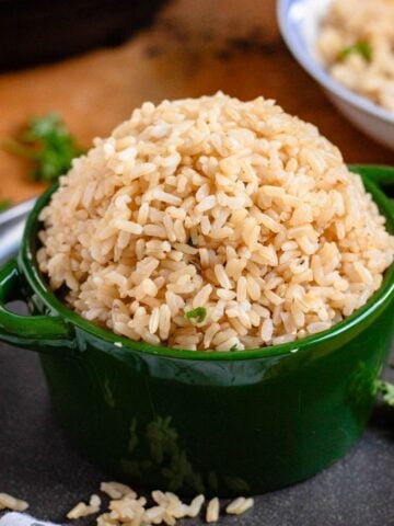 A green bowl filled with cooked brown rice sits on a green bowl with scattered grains and herbs around it.