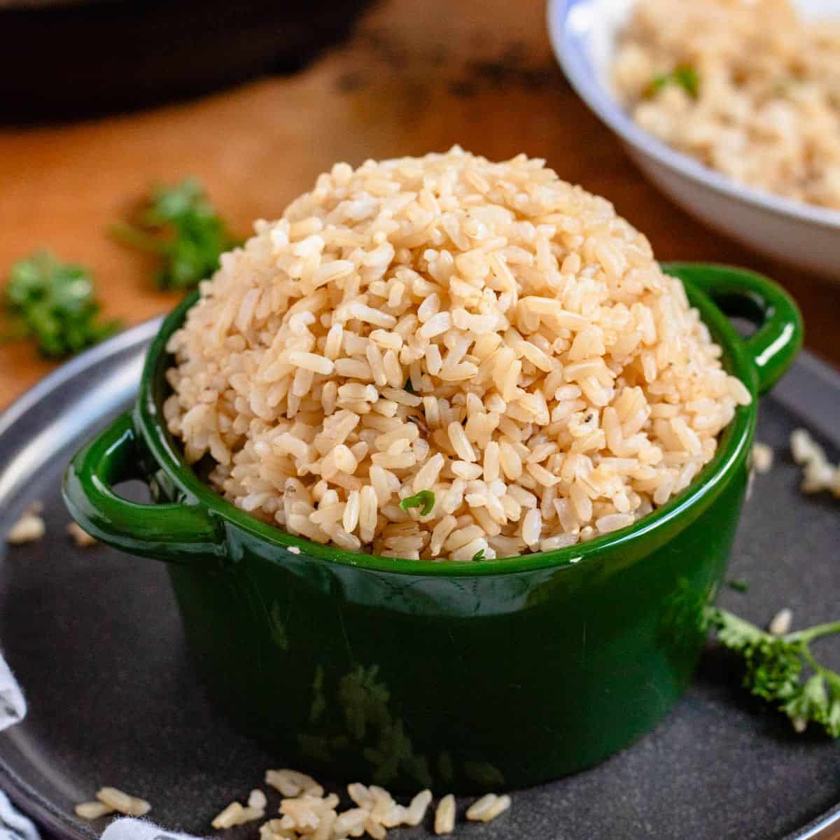A green bowl filled with cooked brown rice sits on a green bowl with scattered grains and herbs around it.