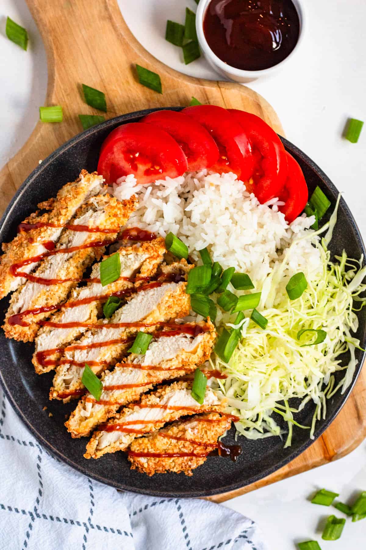 Crispy sliced chicken katsu drizzled with sauce, served over white rice with shredded cabbage, fresh tomato slices, and green onions in a black plate.