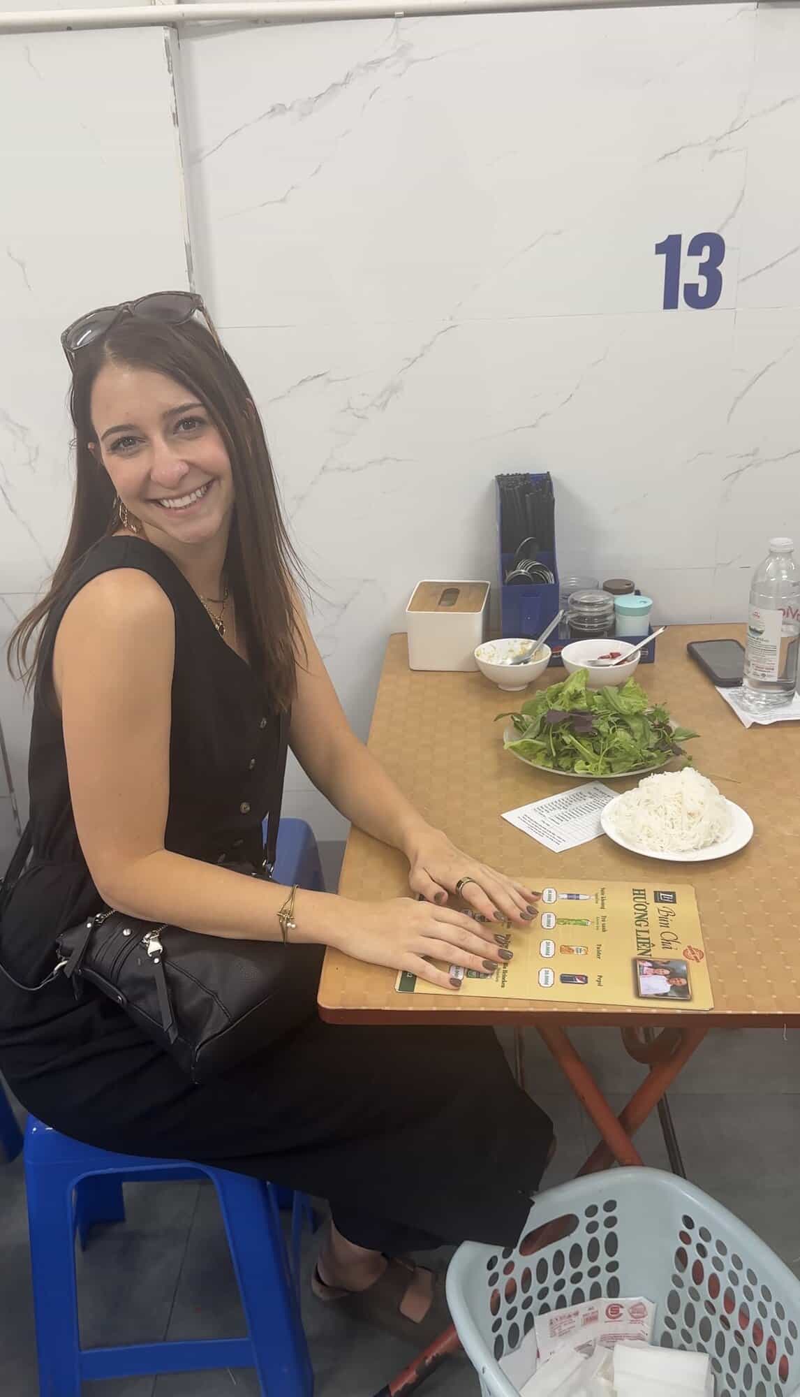 A girl smiling at a dinner table with a pile of herbs and vermicelli noodles.