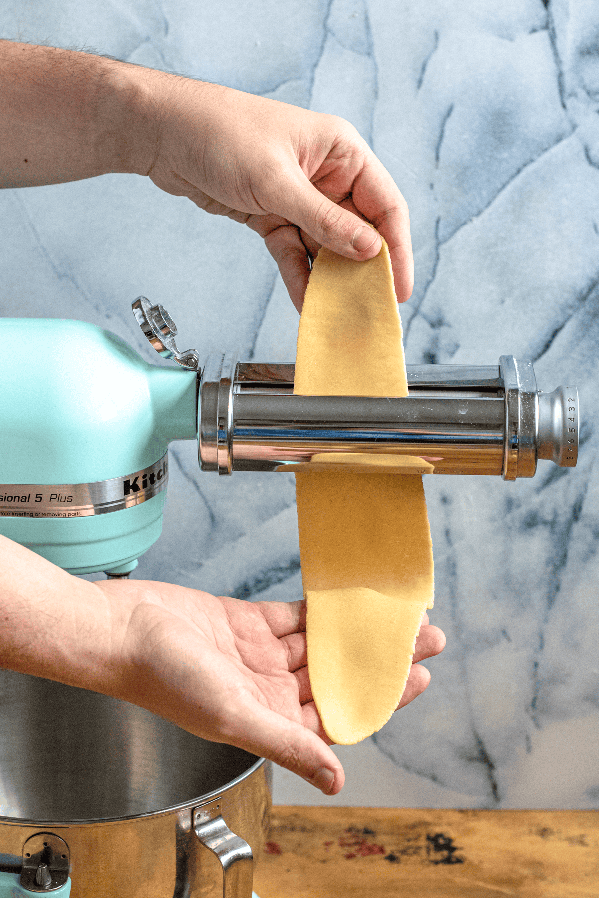 Hands feeding fresh pasta dough through a stand mixer pasta roller attachment to make long sheets of homemade pasta.