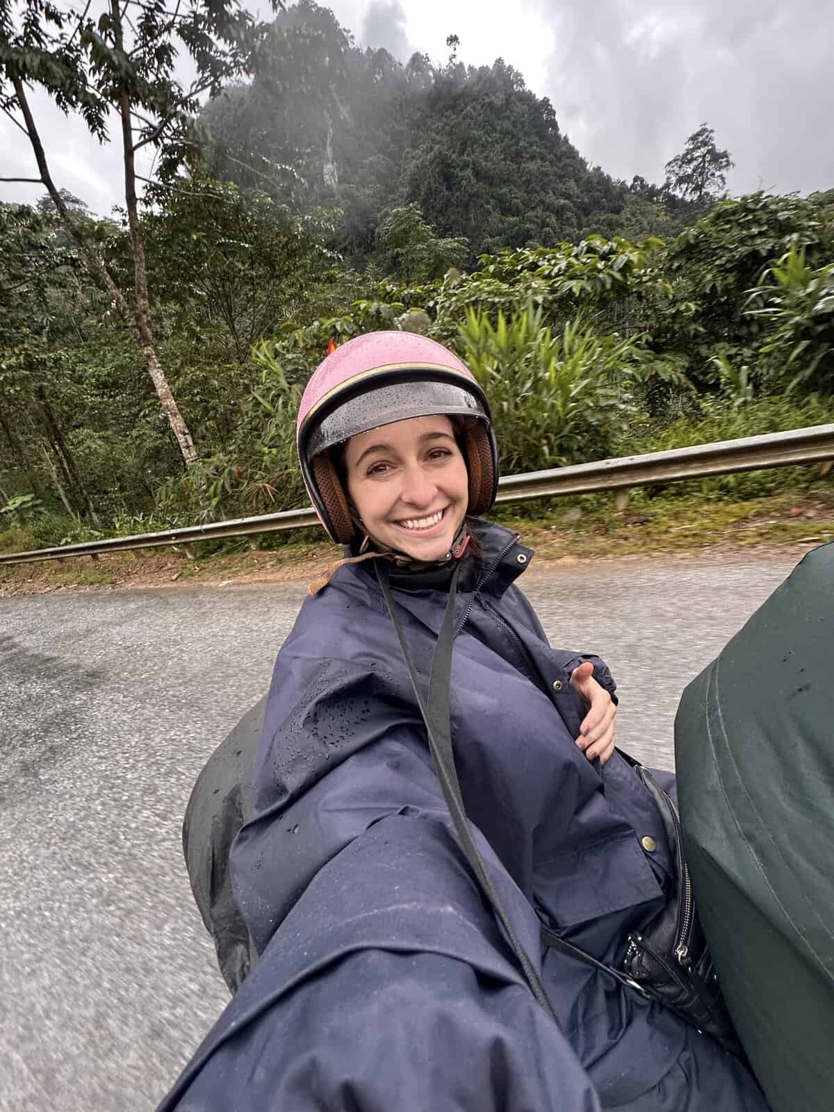 A girl on the back of a motor bike with a pink helmet on taking a selfie.
