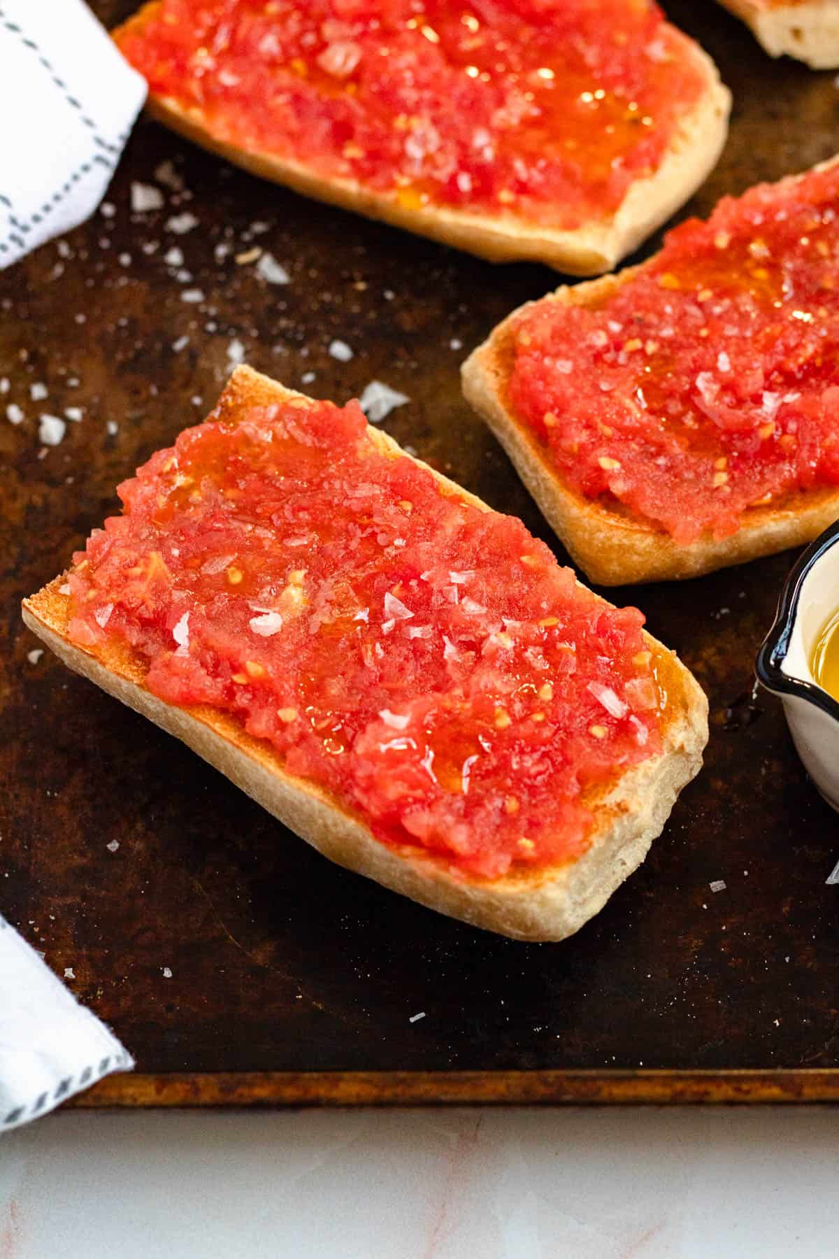 Close-up image of pan con tomate arranged on a baking sheet with a small measuring cup of olive oil.
