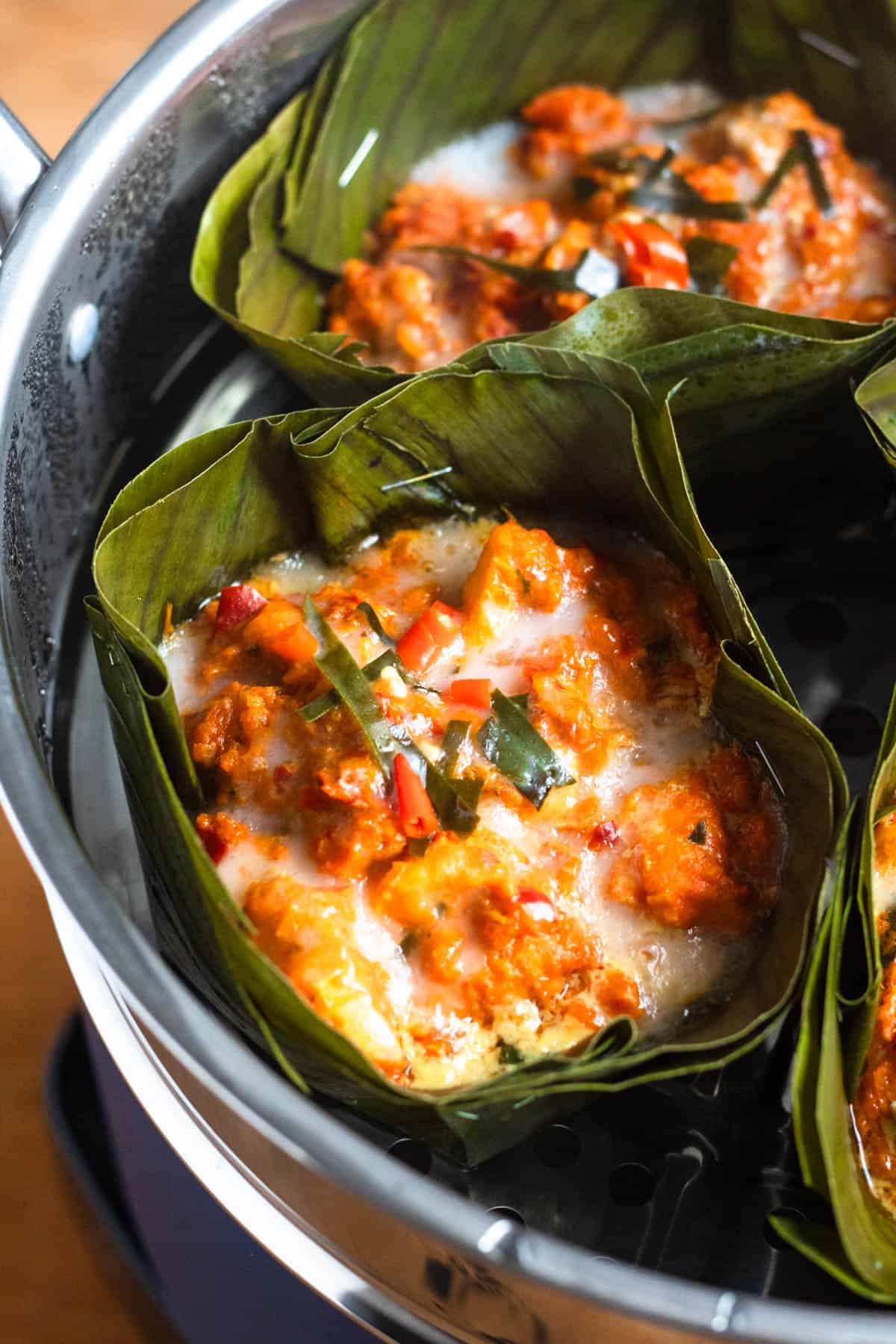 Banana leaf–wrapped amok curry steaming in a steamer, topped with coconut milk and chili peppers.