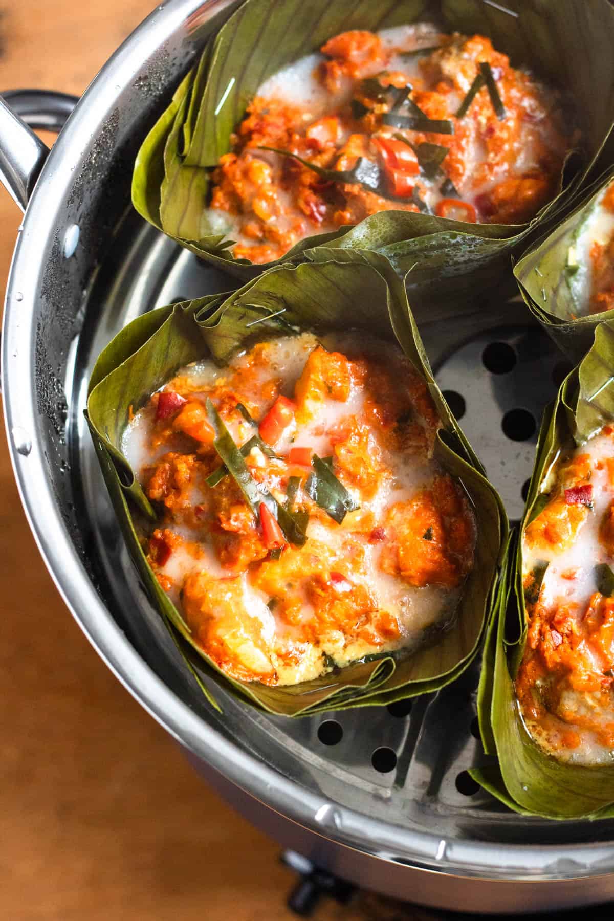 Rich banana leaf–wrapped Cambodian Amok Curry steaming in a steamer, topped with coconut milk and chili peppers.