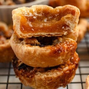 Stacked Authentic Canadian Butter Tarts with flaky crusts and gooey caramel filling, one with a bite taken out, on a cooling rack.