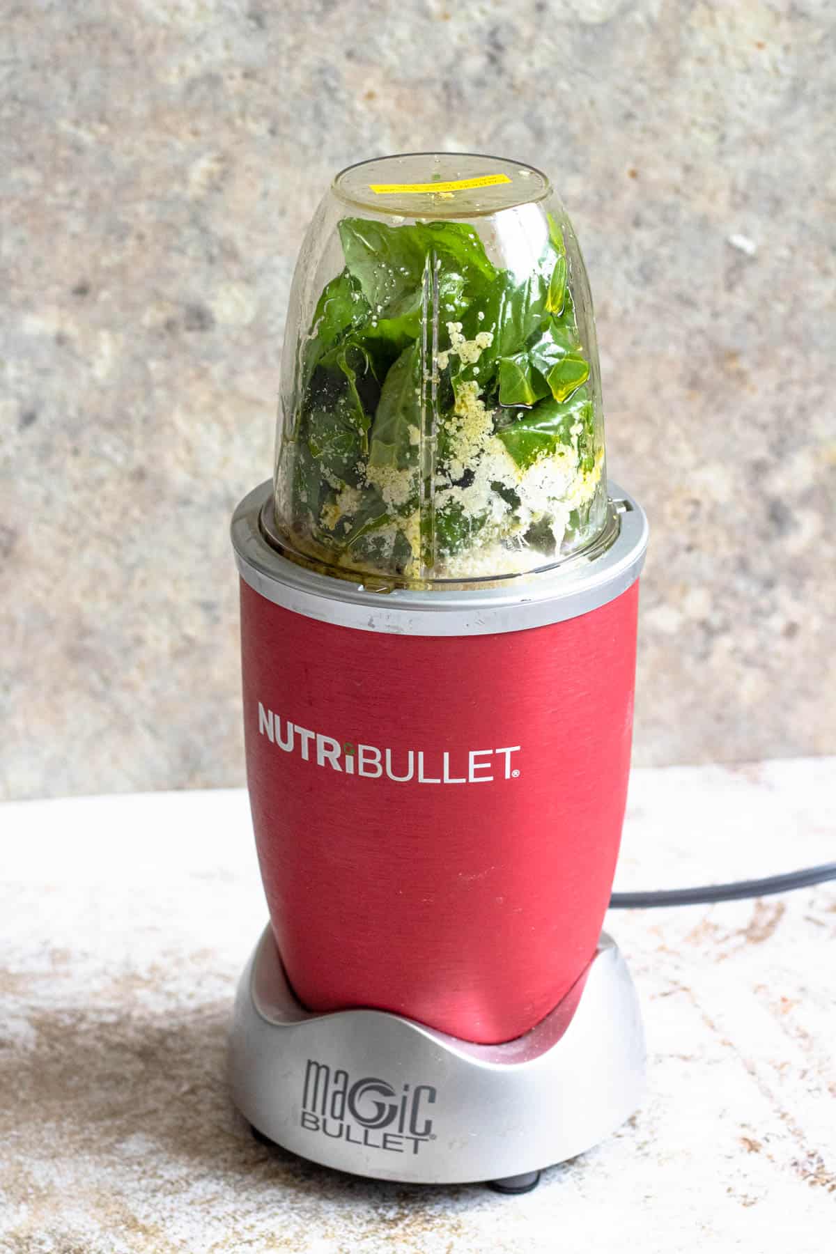Blender cup filled with fresh basil, cheese, and ingredients for pesto.