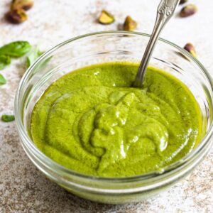 Creamy Pistachio Pesto in a glass bowl with a spoon, surrounded by pistachios and basil.