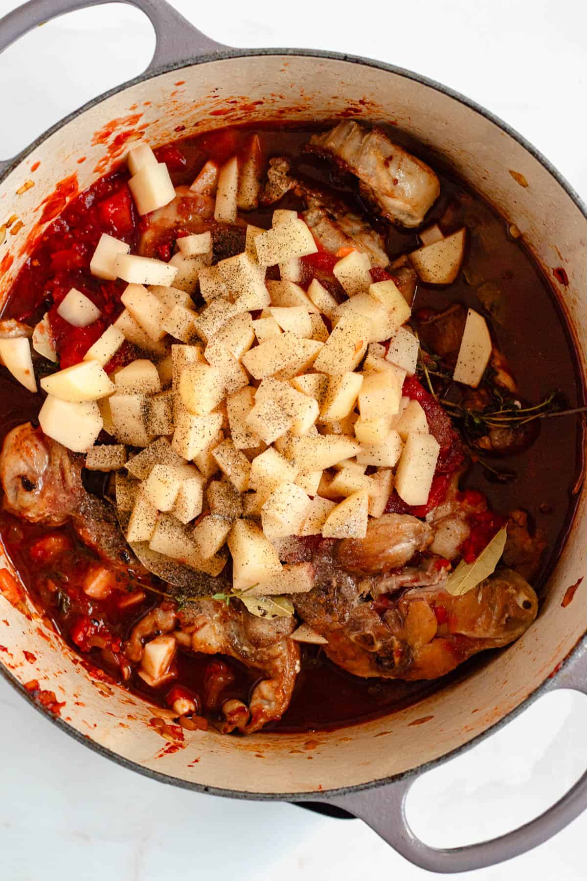Red wine and crushed tomatoes poured into the pot with potatoes, salt, and pepper added around the rabbit pieces.
