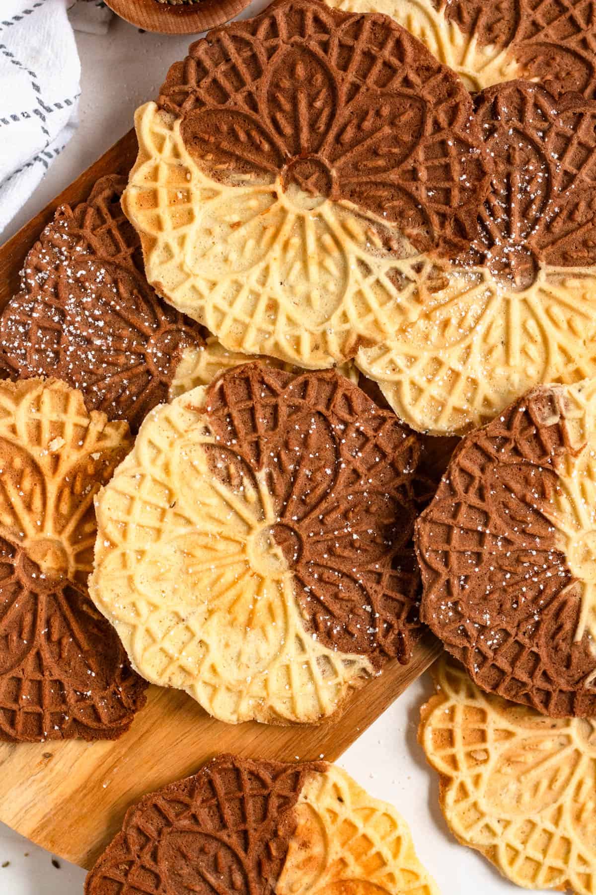 Vanilla and chocolate Swirl Pizzelle cookies lightly dusted with powdered sugar and stacked on a wooden board.
