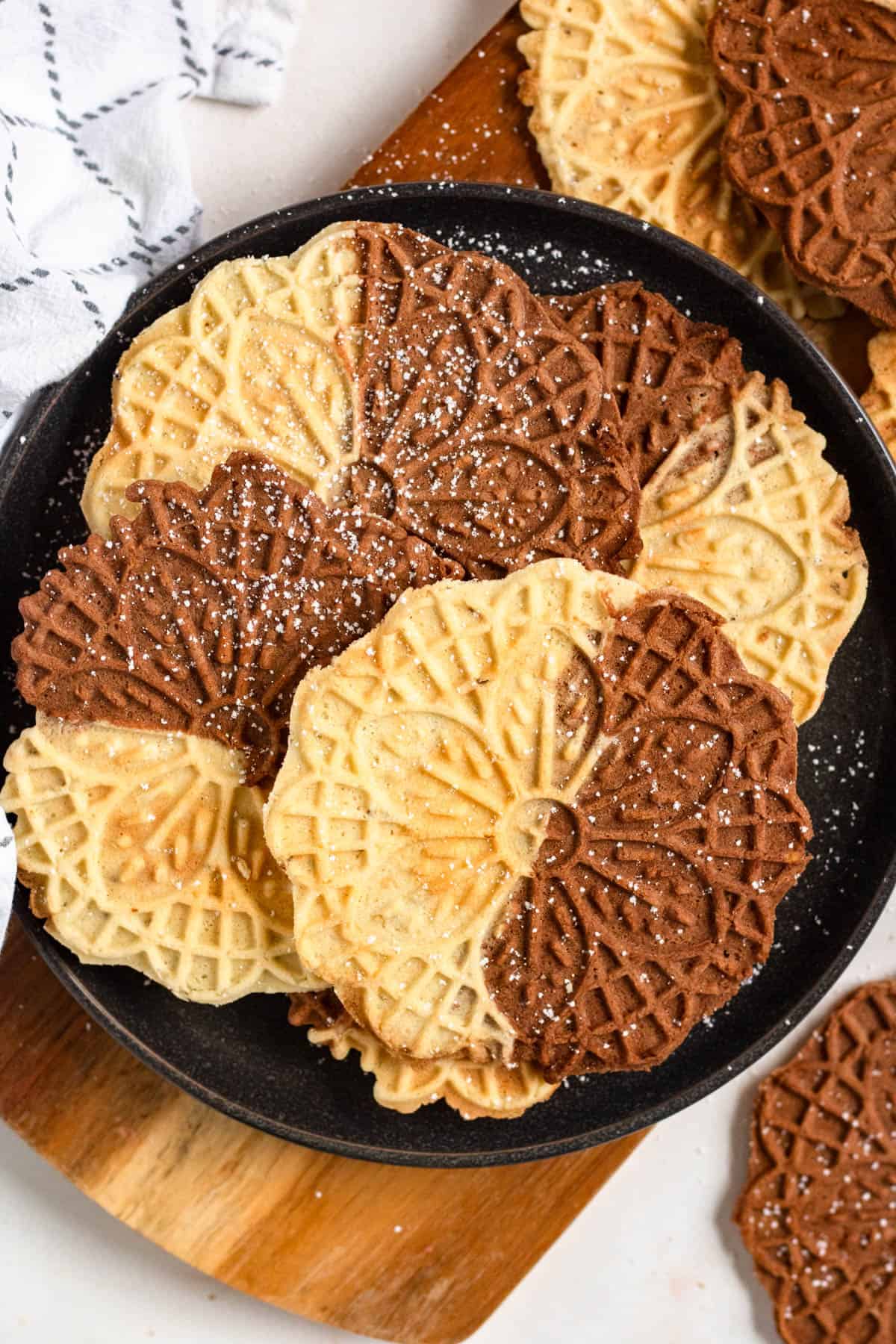 Chocolate and Vanilla Swirl Pizzelle cookies stacked on a black plate and dusted with powdered sugar.