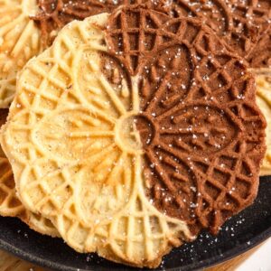 Vanilla and Chocolate Swirl Pizzelle cookies stacked on a black plate and dusted with powdered sugar.