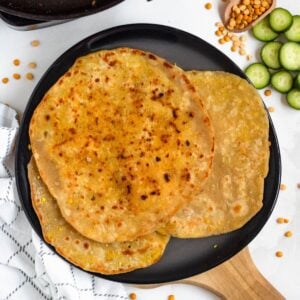 Golden-brown Dhal Puri (Soft Split Pea Flatbread) stacked on a black plate, with split peas and sliced cucumbers on the side.