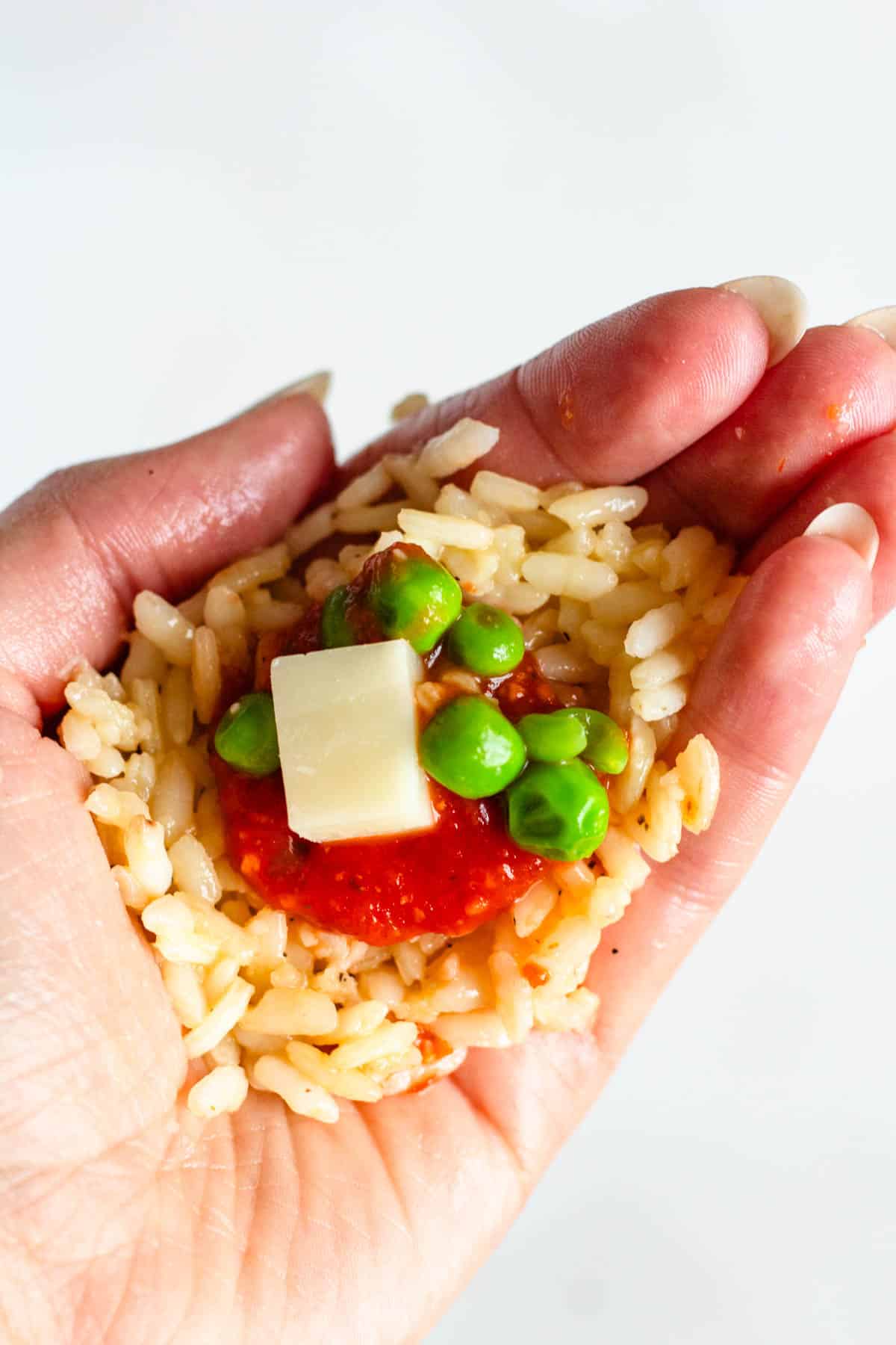Cooked rice in a hand filled with sauce, green peas, and a cube of mozzarella cheese.